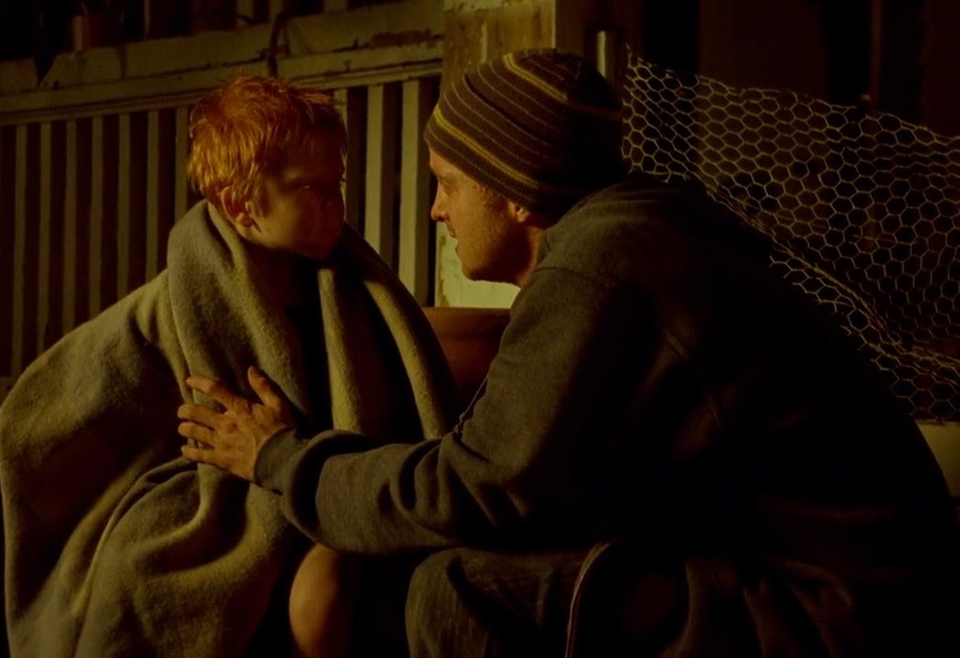 A haggered Jesse (on the left) wraps a blanket around the small red--haired boy in order to comfort him and keep him warm as he waits outside a dilapidated house. The boy is dirty and neglected.